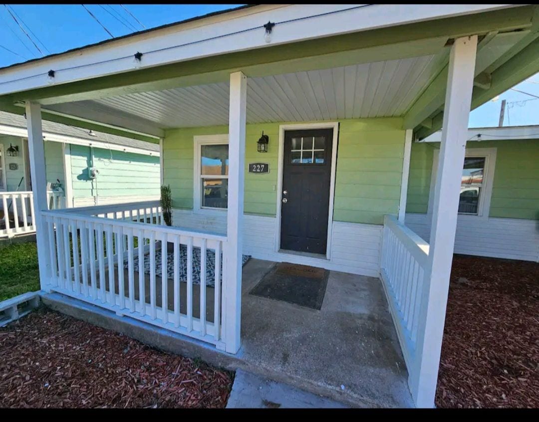 The front porch is framed by white railings, featuring a simple welcome mat positioned at the entrance. Light green siding contrasts with the dark front door. Small shrubs and decorative stones create a tidy appearance in the yard.