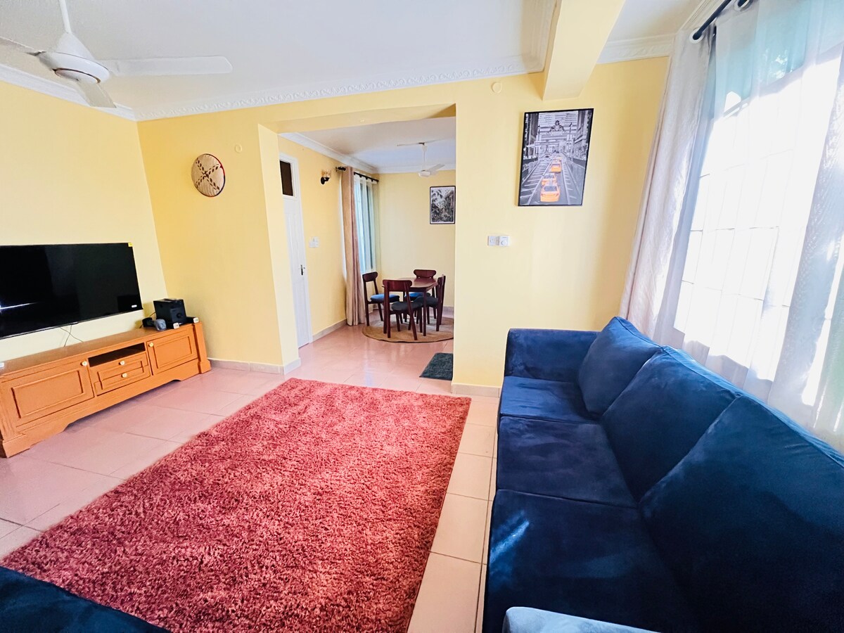 An inviting living area features a large dark blue sectional sofa and a red area rug on tiled flooring. A wall-mounted television sits on a wooden unit. Light enters through large windows covered by sheer curtains, leading to a dining area with a table and chairs.