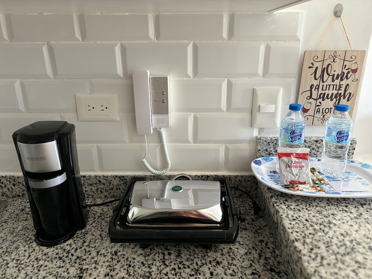 A kitchen counter is pictured, featuring a coffee maker, a hot plate, and a phone mounted on the wall. Bottled water is placed on a tray alongside small snacks. The counter is made of light-colored granite, with a textured white backsplash adding a clean aesthetic.