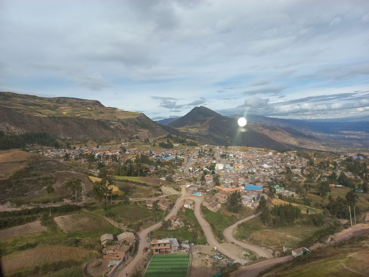 A panoramic view of a valley reveals a small town nestled among rolling hills. Green fields and patches of cultivated land are visible, along with a clear sky stretching out above. The winding road leads through the landscape, enhancing the sense of tranquility.