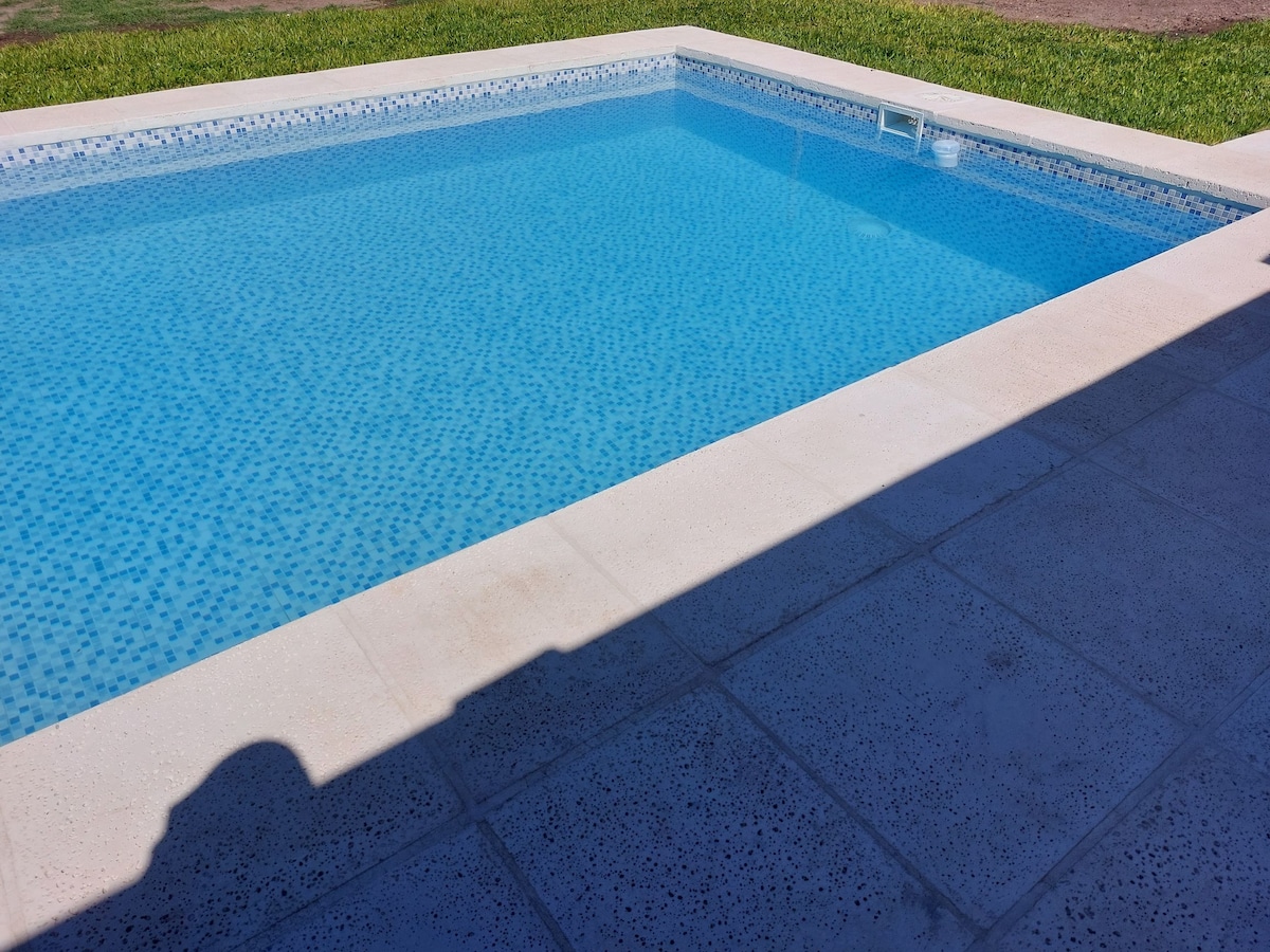 A rectangular swimming pool features clear blue water, bordered by light-colored tiles. The pool is surrounded by a grassy area, with a shadow indicating the presence of a nearby structure. The scene conveys a tranquil outdoor environment ideal for relaxation.