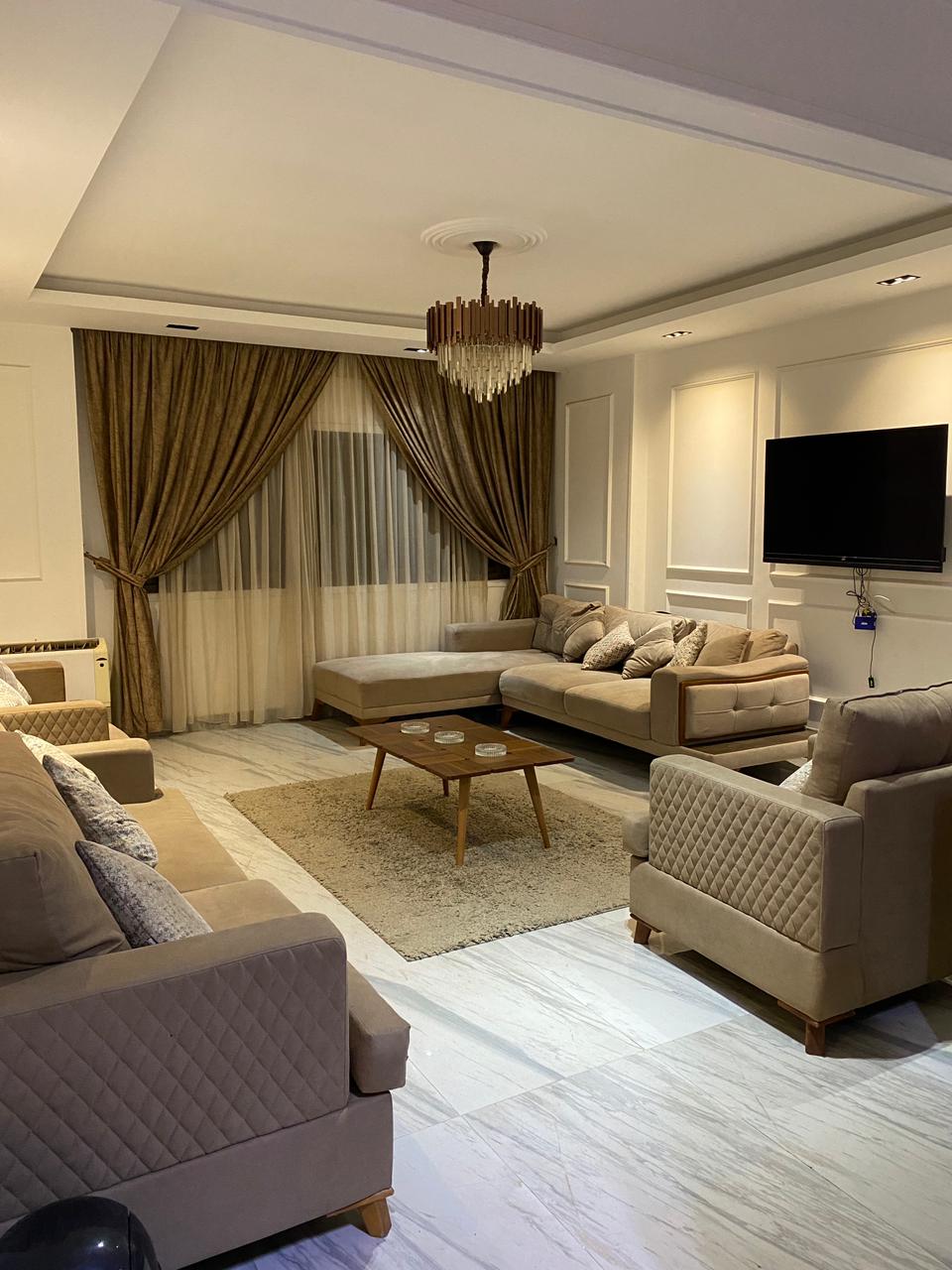A comfortable living area is presented with plush sofas and armchairs, arranged around a wooden coffee table. Soft lighting highlights the room, while elegant drapes frame the windows. A wall-mounted television is unobtrusively placed, adding functionality to the inviting space.
