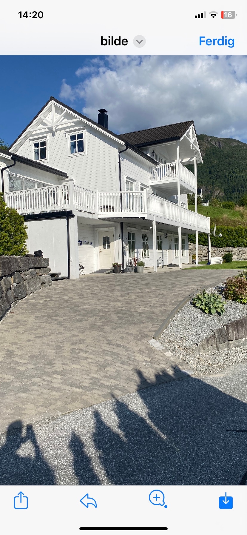 A two-story white house is seen with a spacious front driveway made of pavers. The property features a balcony on the upper level, surrounded by a landscaped area with grass and decorative stones, with mountains in the background under a clear blue sky.