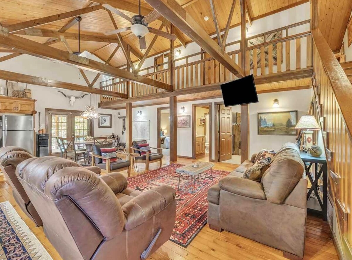 An open-concept living area features a high ceiling supported by wooden beams. Comfortable seating is arranged around a central coffee table on a patterned area rug. Natural light streams in through large windows, illuminating the inviting space with a warm ambiance.