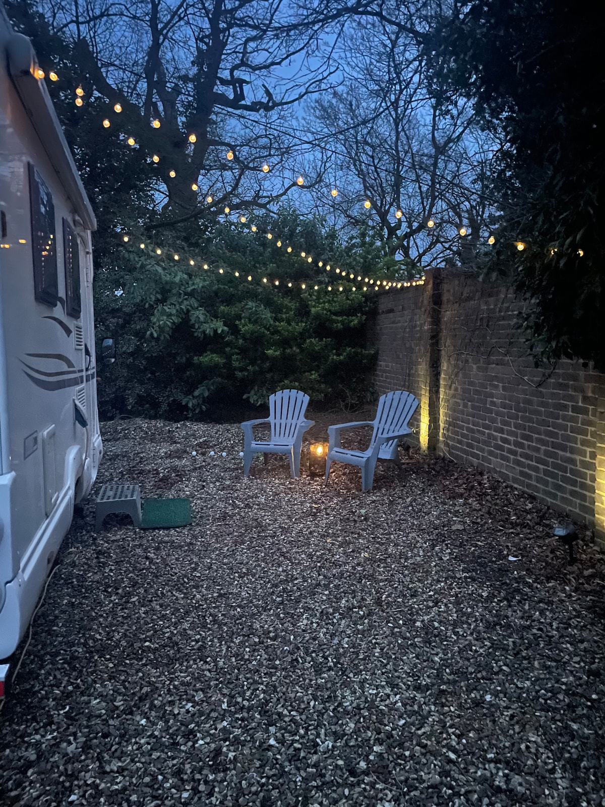 A cozy outdoor area is featured beside the motorhome, with two outdoor chairs positioned around a small fire. Soft string lights hang overhead, creating a warm ambiance amidst the surrounding greenery and a brick wall, set against the twilight sky.
