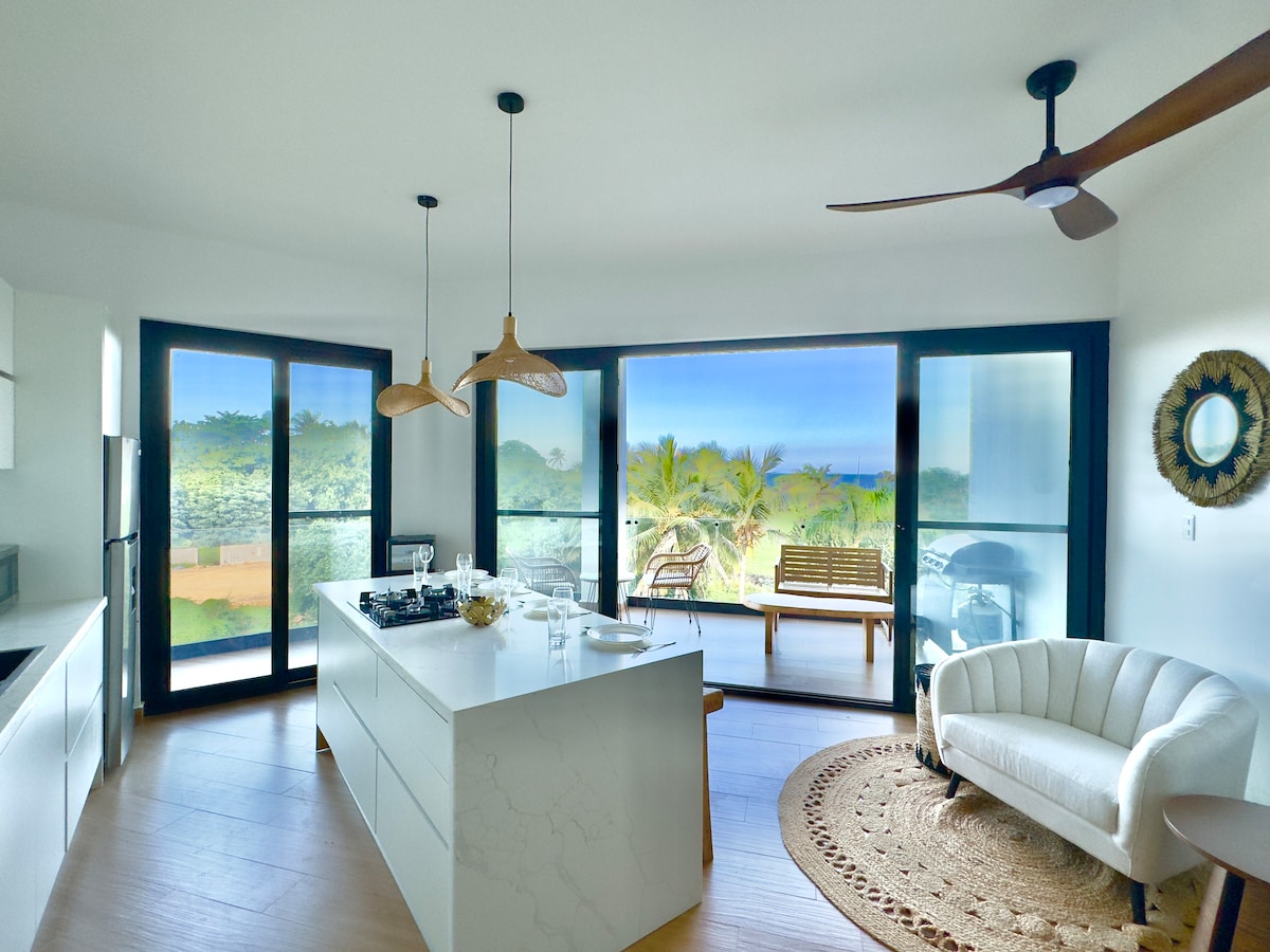 A bright and airy kitchen is visible, featuring a spacious island with a stove and minimalist decor. Floor-to-ceiling glass doors open to a balcony, revealing views of lush greenery and the ocean. A cozy seating area is set with a curved couch and woven rug.