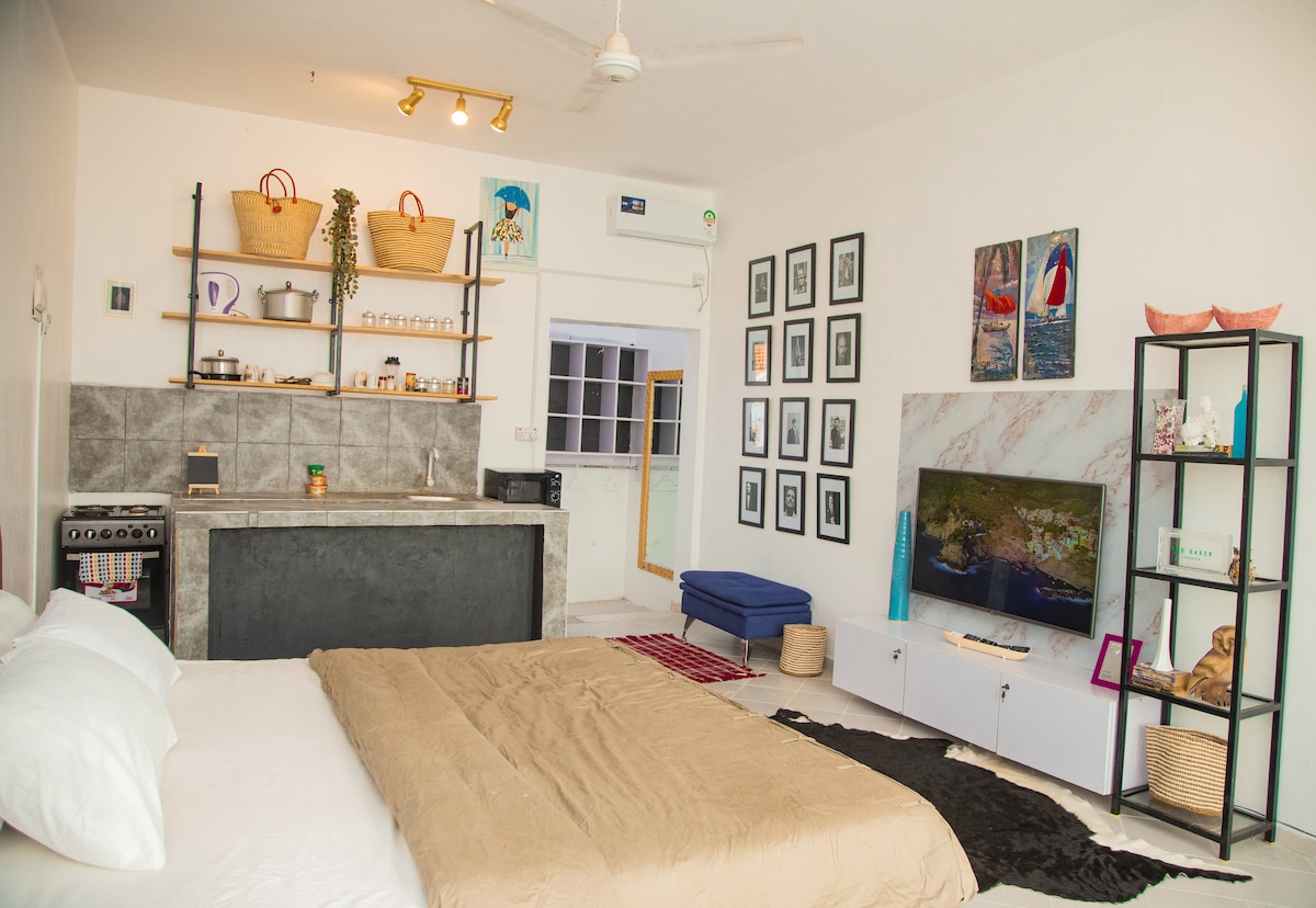 A spacious room features a large bed with neutral bedding, adjacent to a kitchenette equipped with shelves displaying various items. A sleek entertainment unit holds a smart TV, while art prints adorn the walls. Light streams in from a nearby window, enhancing the airy atmosphere.