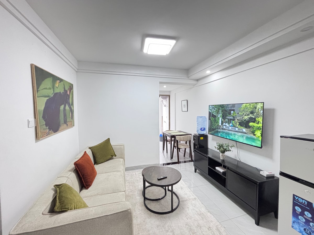 The living room showcases a neutral-toned couch with accent pillows. A large coffee table is positioned in front, complementing the seating area. A 55” Smart TV is mounted above a spacious TV stand, with a small dining area visible in the background.