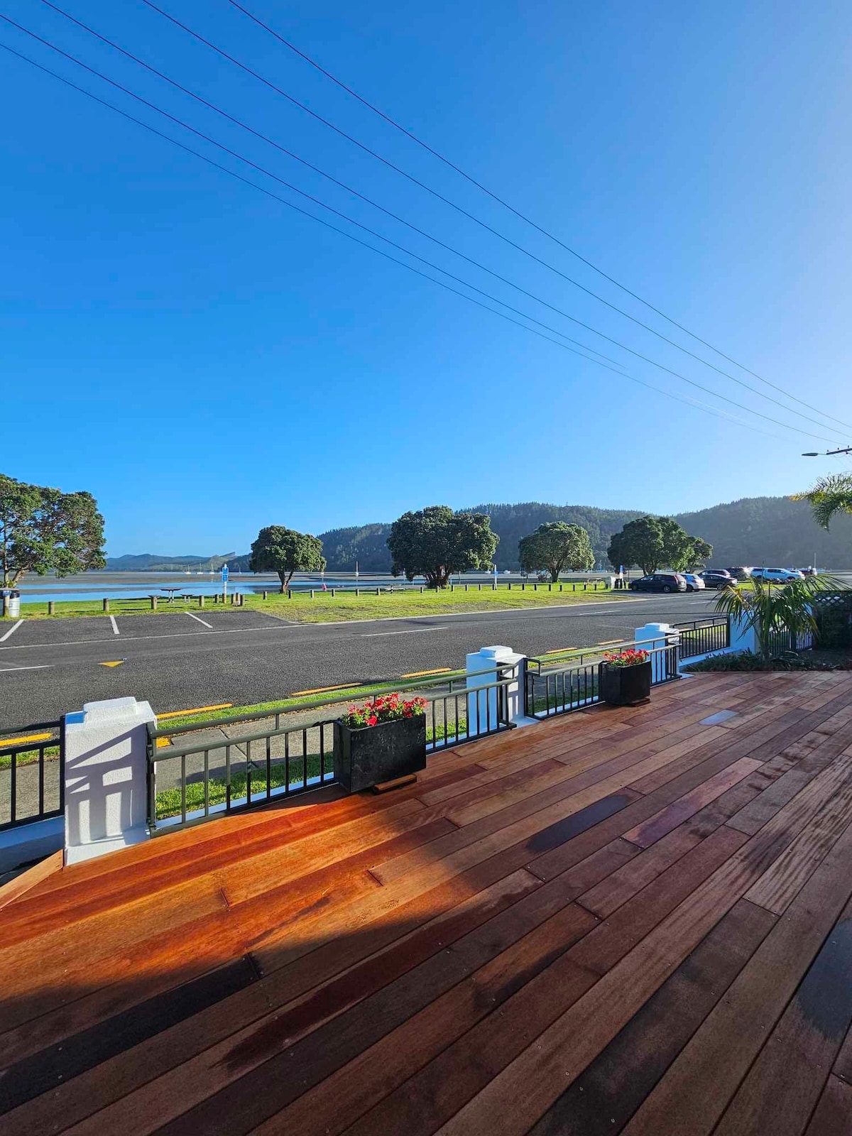 A spacious wooden deck is visible, offering a clear view of the surrounding landscape. Lush green hills are in the background, while the foreground features a well-maintained lawn and flowering planters. Blue skies dominate the scene, creating a relaxing outdoor space.