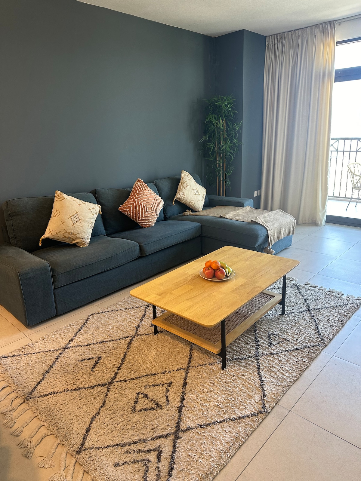 A cozy living area features a dark sofa adorned with decorative pillows. A wooden coffee table is positioned at the center, holding a bowl of fruit. Light-colored curtains frame a balcony door, while a potted plant adds a touch of greenery.