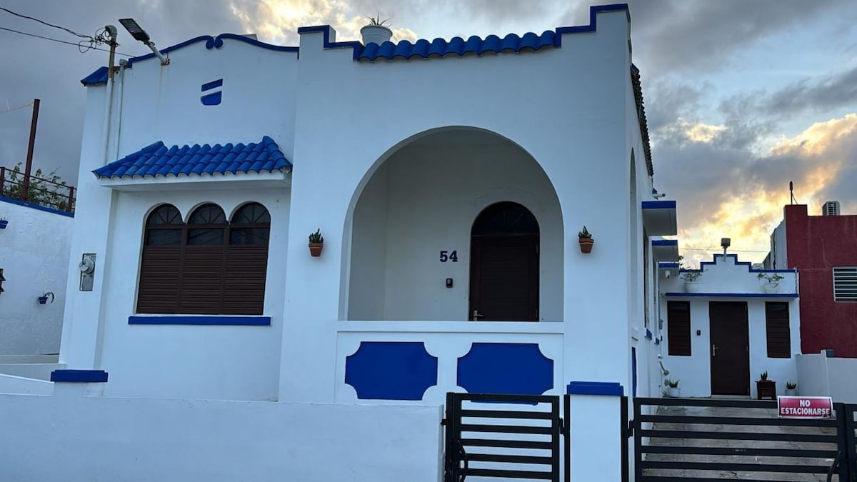 A charming white building with blue accents features arched windows and a decorative archway at the entrance. Potted plants adorn the exterior, and a secure gate leads to the property. The sky is partly cloudy, enhancing the serene atmosphere.