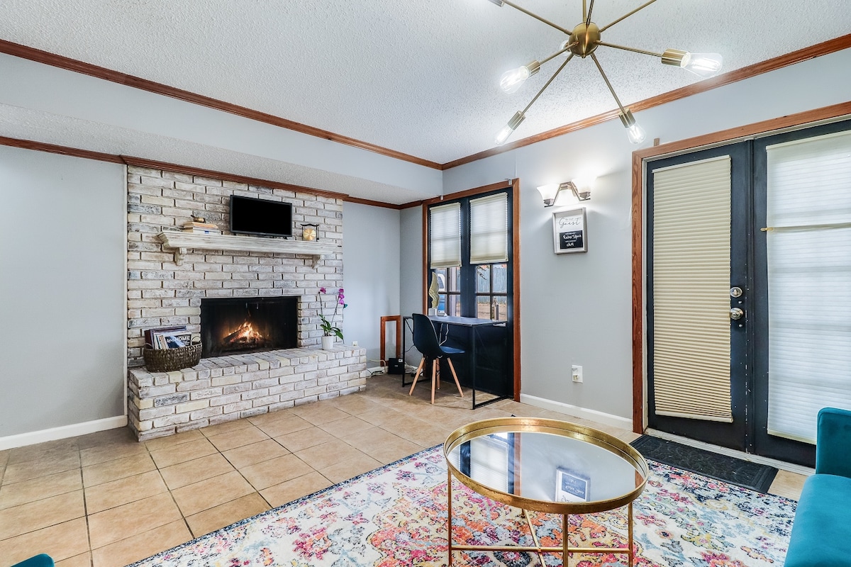 The living area features a stone fireplace, providing a focal point for relaxation. Natural light filters through the windows, highlighting the colorful area rug beneath a glass-top coffee table. A stylish chair and modern light fixture add contemporary touches to the space.
