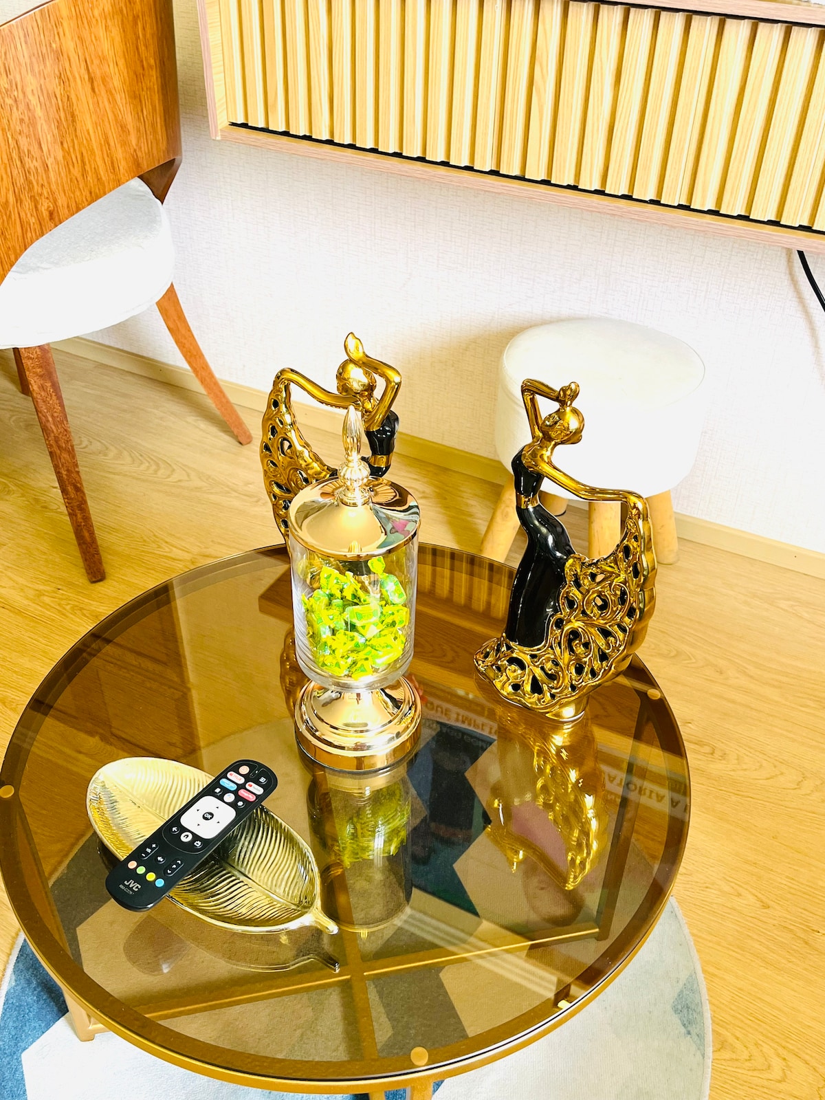 A round glass table is showcased in the living area, featuring a decorative centerpiece with two elegant figurines and a glass container filled with colorful items. A remote control sits beside a stylish metallic dish, complemented by a wooden chair in the background.