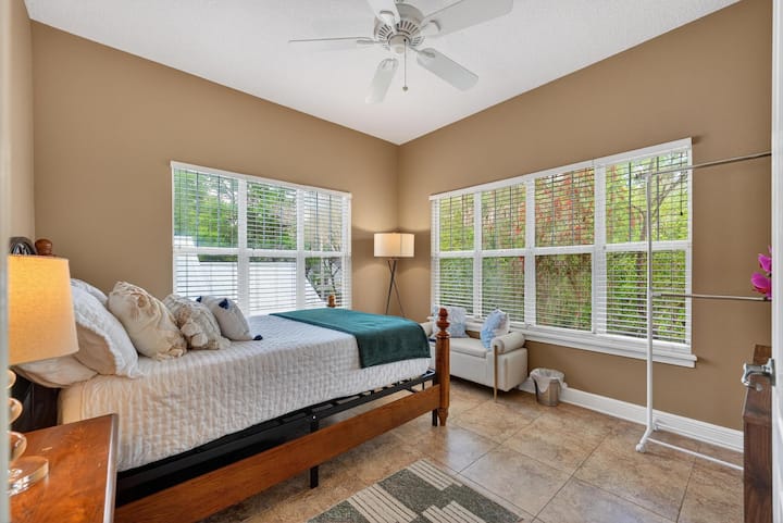 Um quarto iluminado com teto alto, janelas panorâmicas com vista para o jardim, uma cama queen-size confortável com toques em estilo boho e um banco de leitura aconchegante ao lado da janela. Relaxe em um espaço ensolarado com iluminação ambiente aconchegante.
