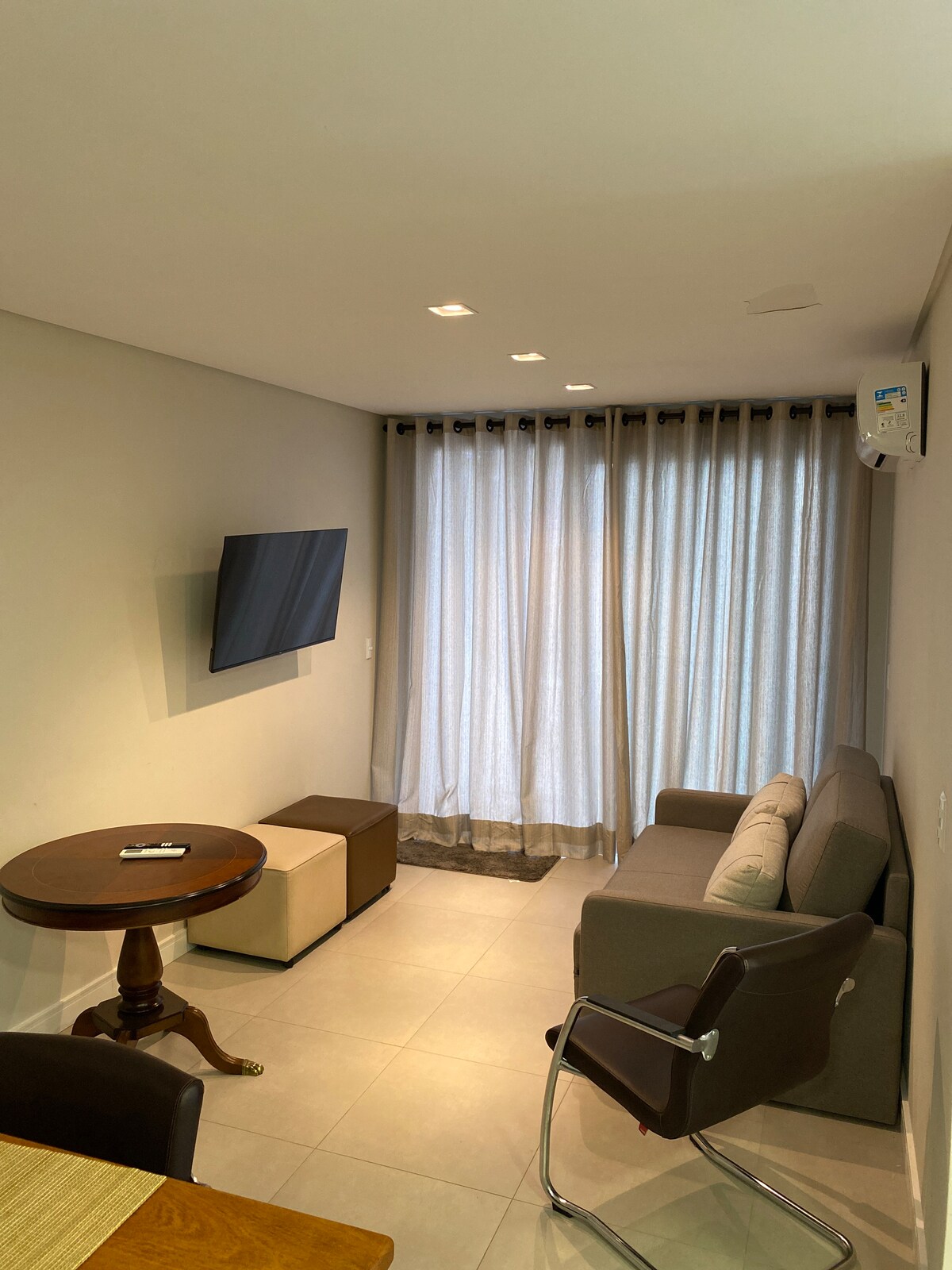 A modern living area features a sleek sofa accompanied by a small coffee table. A flat-screen TV is mounted on the wall opposite large curtains that filter natural light. Neutral tones create a relaxed atmosphere, enhancing comfort in the space.