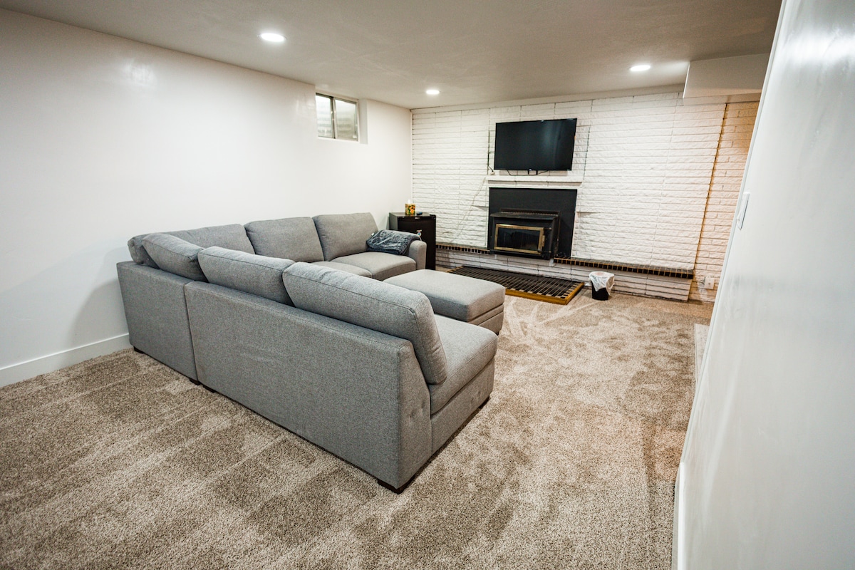 A spacious basement lounge area features a large sectional sofa arranged for relaxation and enjoyment. A mounted television is positioned above a traditional fireplace, providing a cozy focal point. Soft, neutral carpeting enhances the inviting atmosphere of the space.