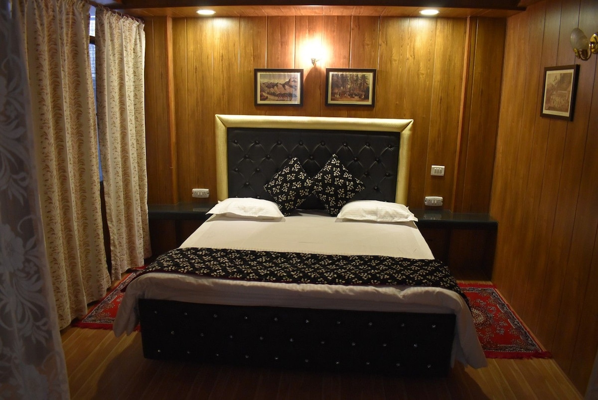 A cozy bedroom is highlighted with wooden paneling and ambient lighting. A spacious bed is centered against the wall, adorned with decorative pillows. The room features nightstands and artwork, contributing to the inviting decor without overwhelming the space.