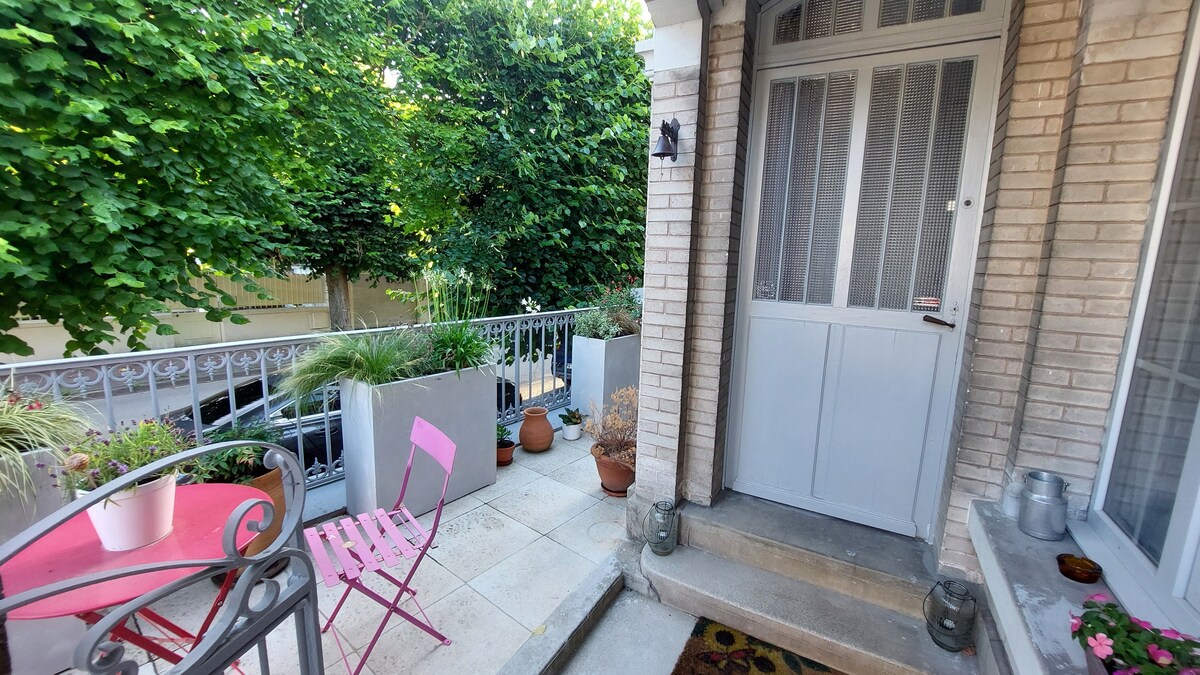 A welcoming entrance area features a small, pink bistro table and a matching chair positioned on a stone patio. Flower pots are arranged around the space, complemented by lush greenery from nearby trees, creating a serene atmosphere.