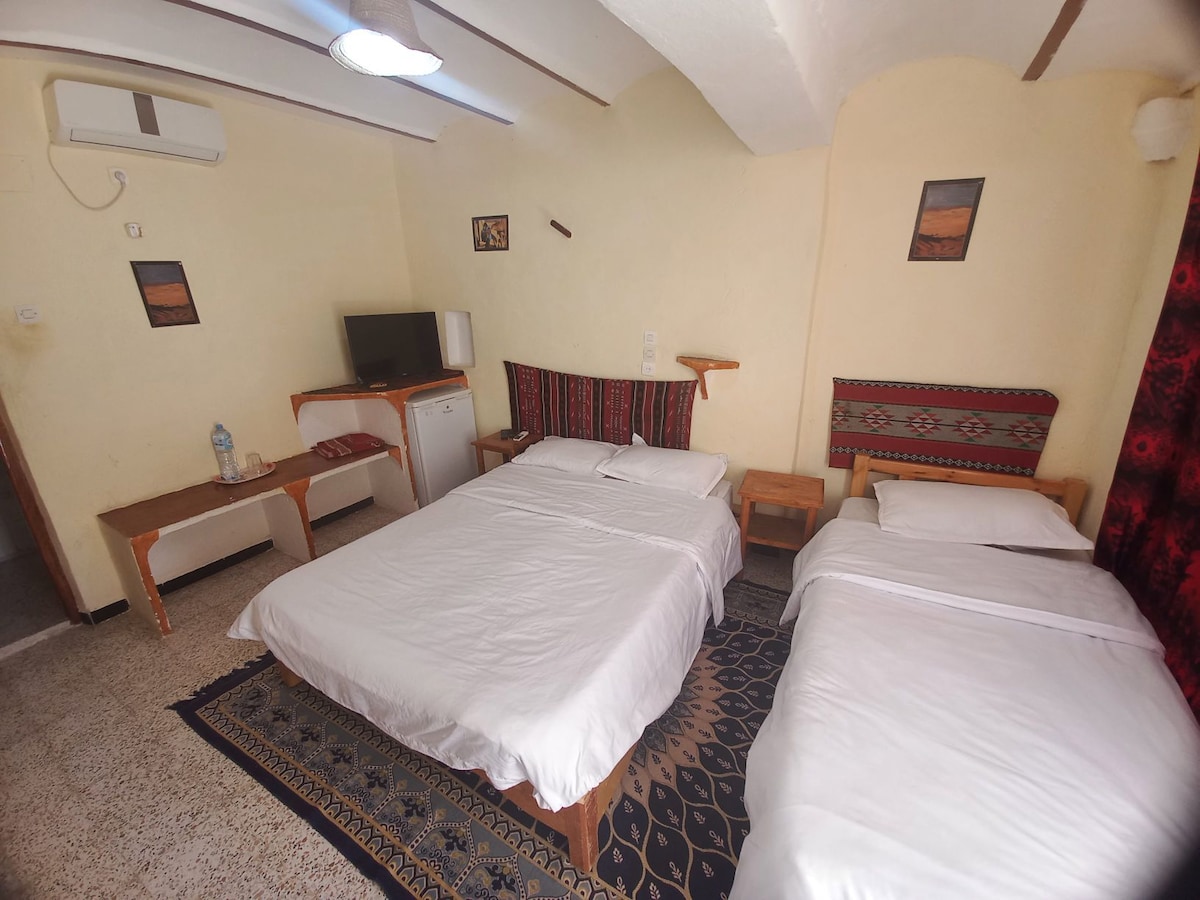 The room features two beds, one spacious and one smaller, both with crisp white linens. A television is positioned on a wooden stand, alongside a mini fridge. Accents of traditional artwork are displayed on the walls, and natural light filters through the window covered by dark curtains.