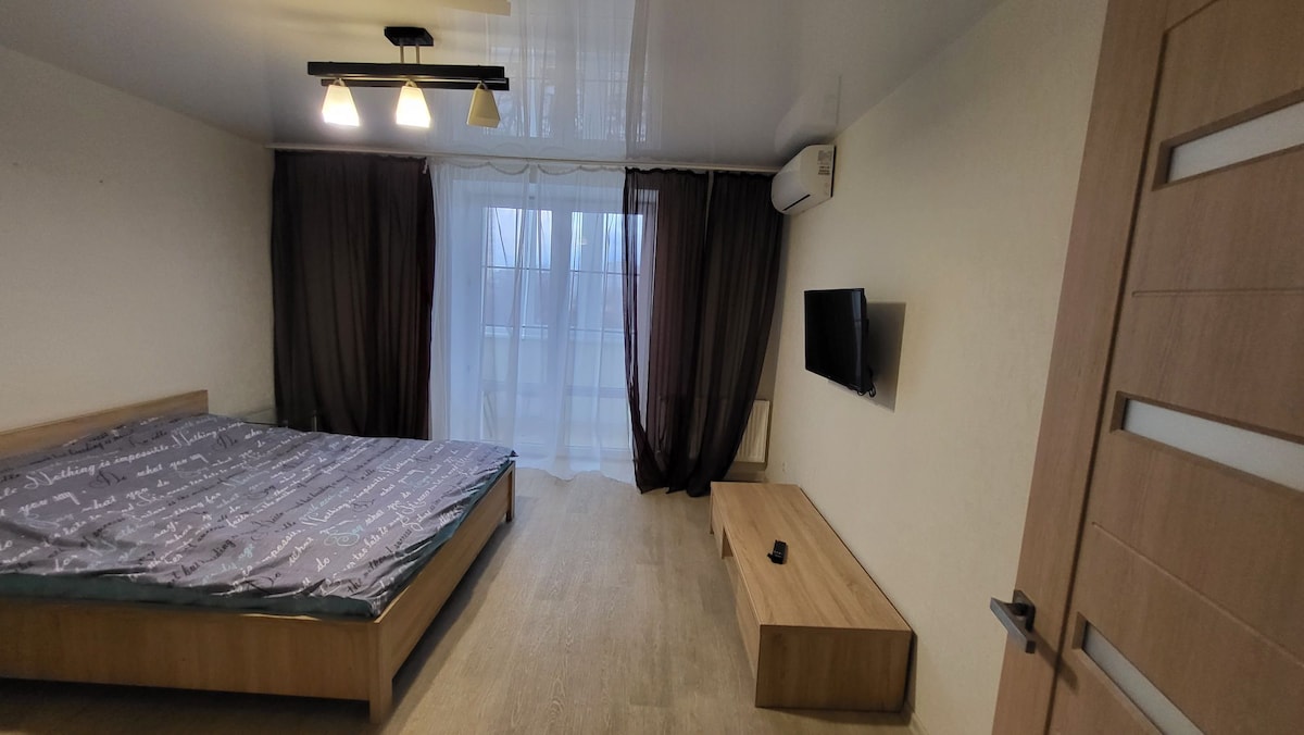 A spacious bedroom features a large bed with a patterned coverlet. Natural light is provided by a window dressed with sheer curtains, while a wooden television stand and flat-screen TV are positioned against a light-colored wall. Dark curtains add warmth to the space.