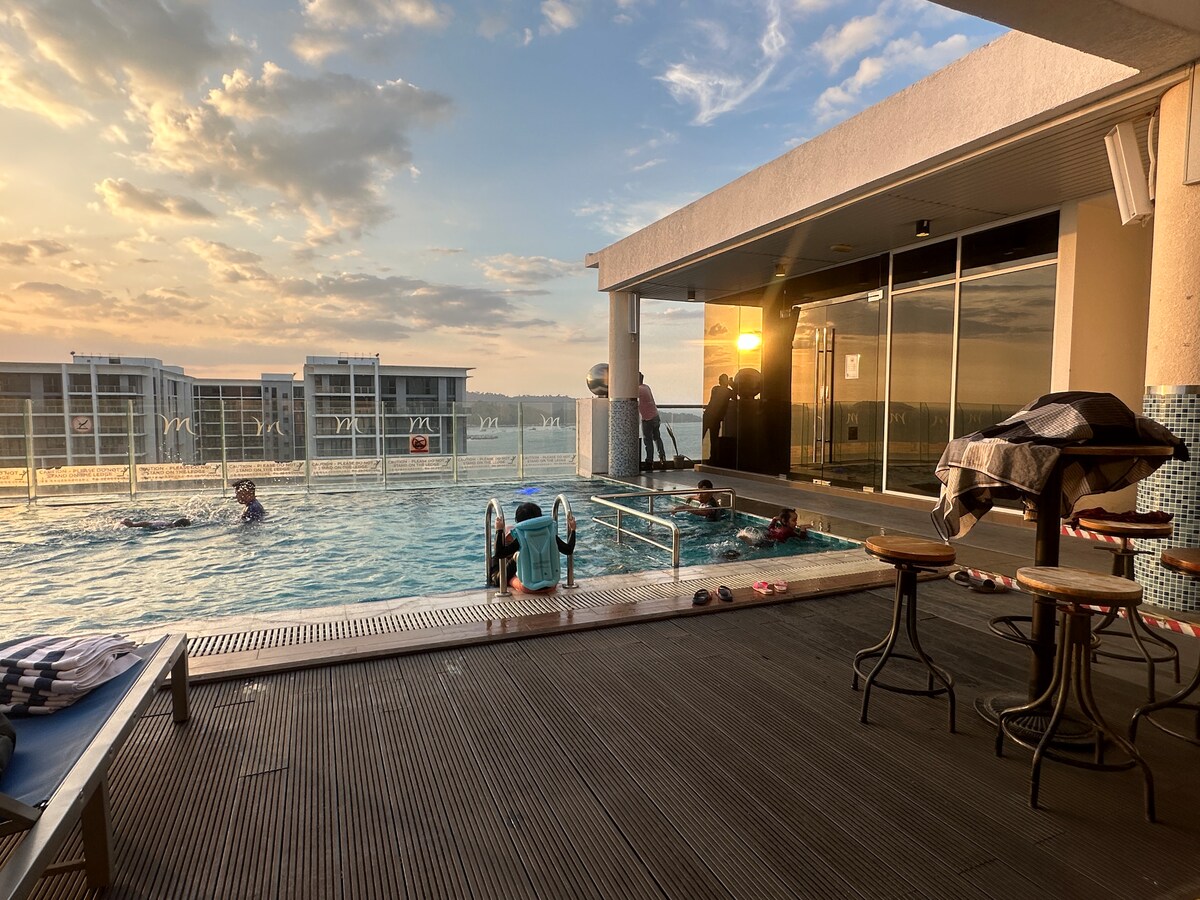 A rooftop swimming pool is depicted, featuring several people enjoying the water as the sun sets in the background. Lounge chairs are positioned nearby, with a small table accompanying them. Panoramic views of the horizon are visible through expansive glass doors.