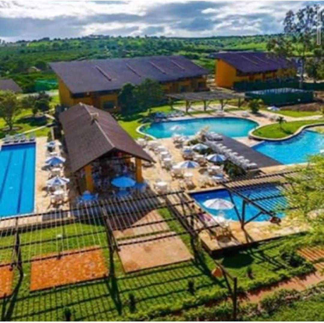 An aerial view showcases multiple swimming pools set amidst lush greenery. Sun loungers with umbrellas are positioned around the pools, while a restaurant structure is seen nearby. Surrounding hills contribute to the tranquil scenery, enhancing the overall inviting atmosphere.
