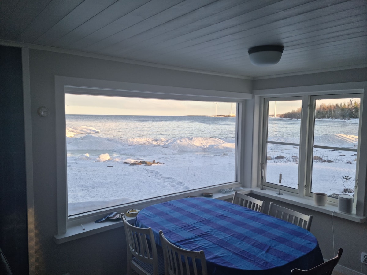 A dining area features a round table with a blue checkered tablecloth, surrounded by white chairs. Large windows offer an unobstructed view of the snowy shoreline, with the sea visible in the distance under a soft, morning light.