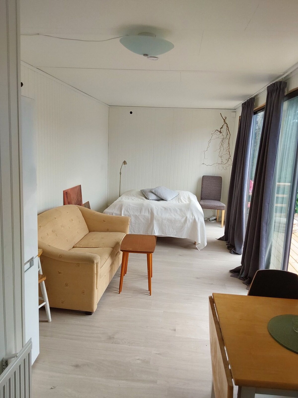 A light-filled living space features a cozy seating area with a beige sofa and a small wooden table. A spacious bed is partially draped with a white cover, and large windows are dressed with soft gray curtains, allowing natural light to enhance the room's ambiance.