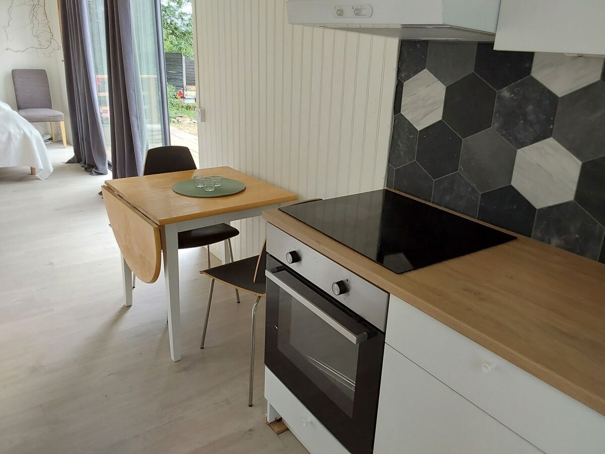 A kitchen space features a modern countertop with a built-in stovetop. A small wooden table with a green placemat is complemented by a black chair. Light-colored cabinetry and a hexagonal tile backsplash enhance the area, while natural light enters through a nearby window.