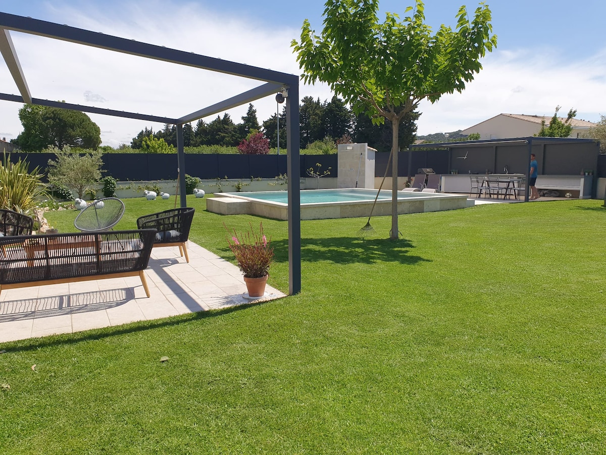 A spacious garden area features a well-maintained lawn, with a covered seating area equipped with comfortable chairs. A swimming pool is visible in the distance, surrounded by decorative shrubbery and trees, creating a relaxing outdoor environment.