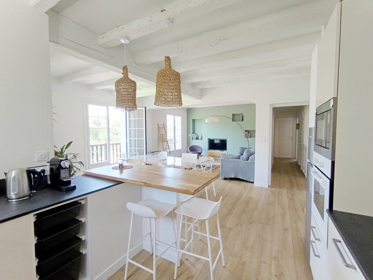 A bright kitchen area features a wooden countertop with two white bar stools and modern appliances. Natural light floods the space through large glass doors leading to a balcony. The adjoining living area can be seen, showcasing comfortable seating and a TV.