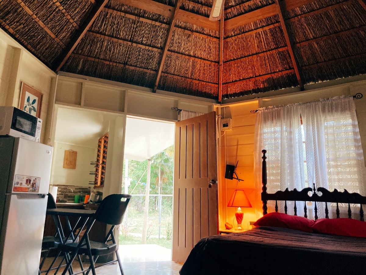 The interior of the thatched hut features a hexagonal design with high, wooden ceilings. A cozy sleeping area includes a bed adorned with a red blanket. A kitchenette and dining area are visible, accompanied by a refrigerator and a microwave. Natural light filters through sheer curtains.