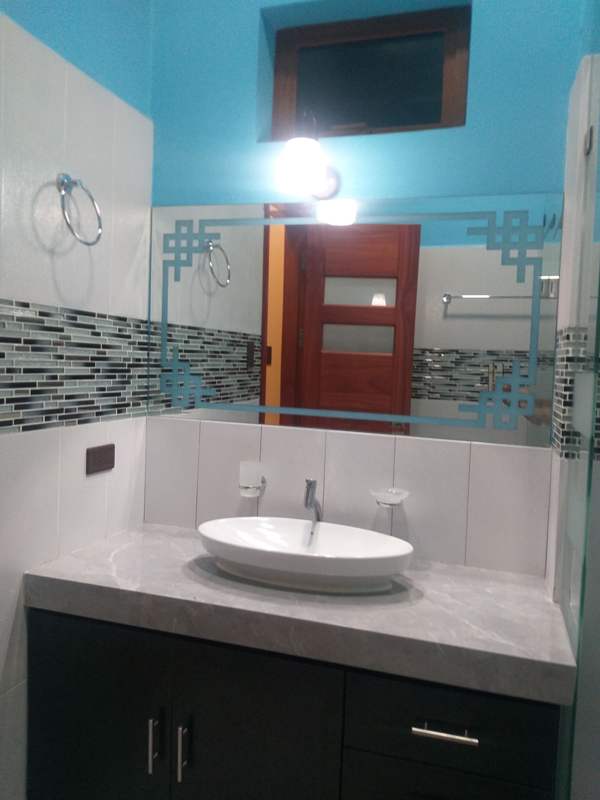 The bathroom features a modern sink with a circular basin set on a gray countertop. A large mirror reflects the blue walls and decorative tile backsplash. Soft lighting illuminates the space, complemented by wooden accents from nearby doors.