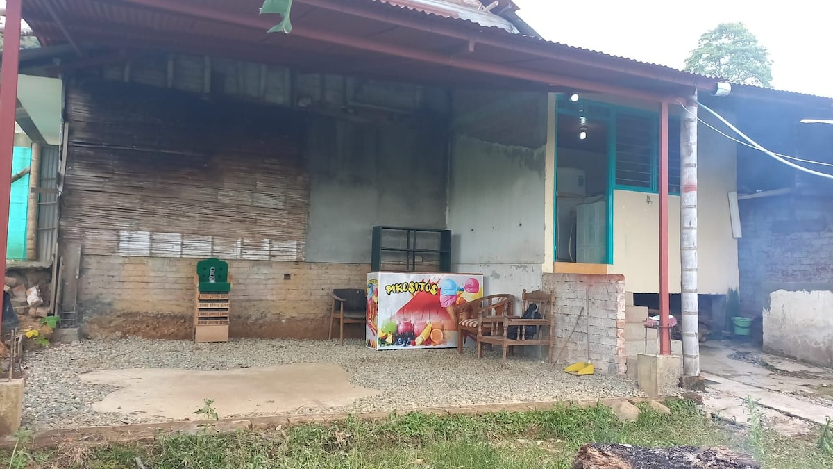 An outdoor area with a gravel surface is visible, featuring a small seating arrangement. A colorful banner is displayed prominently against the wall, while the structure showcases a rustic facade and a mix of materials, reflecting a simple and functional design.