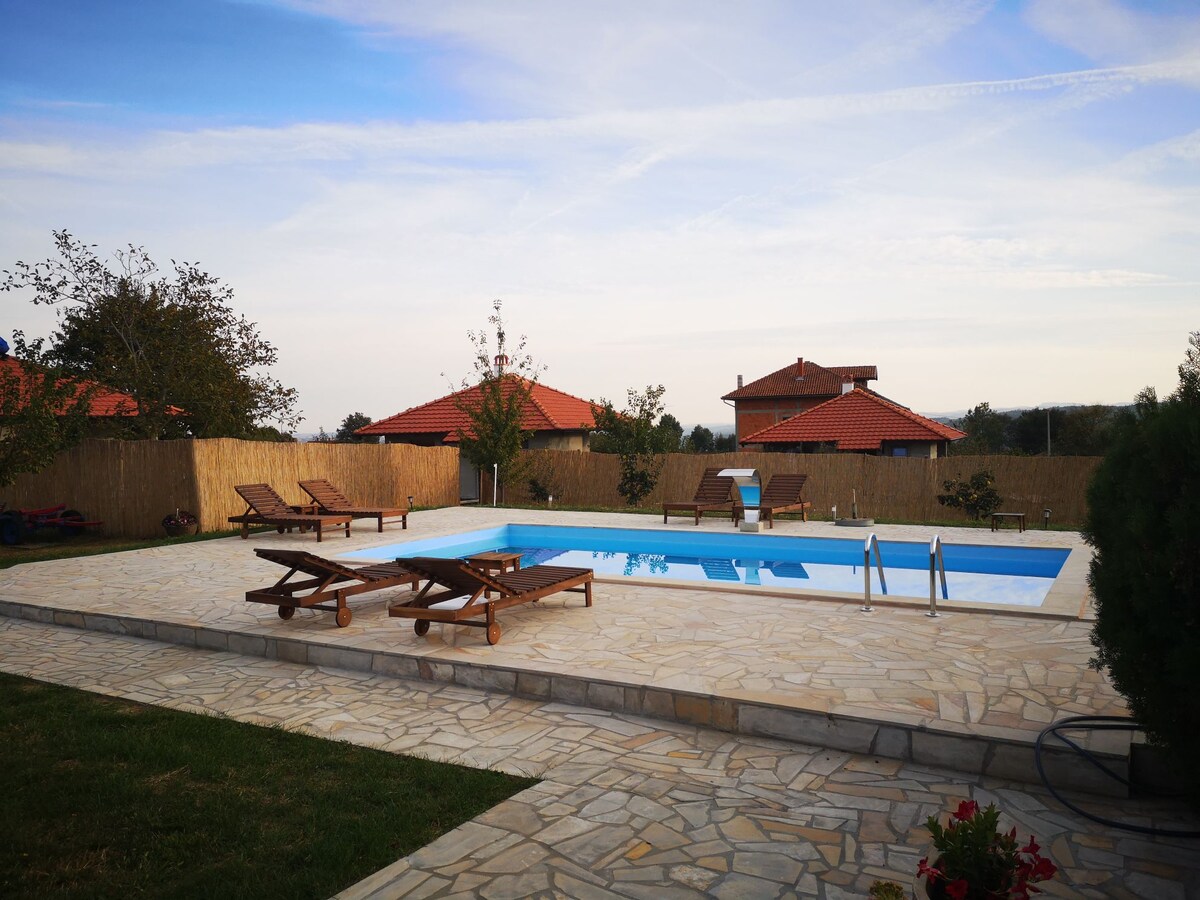 An inviting outdoor space features a spacious swimming pool surrounded by several wooden loungers. Lush greenery and young trees are visible in the background, with two houses featuring red-roofed designs located beyond a wooden fence.