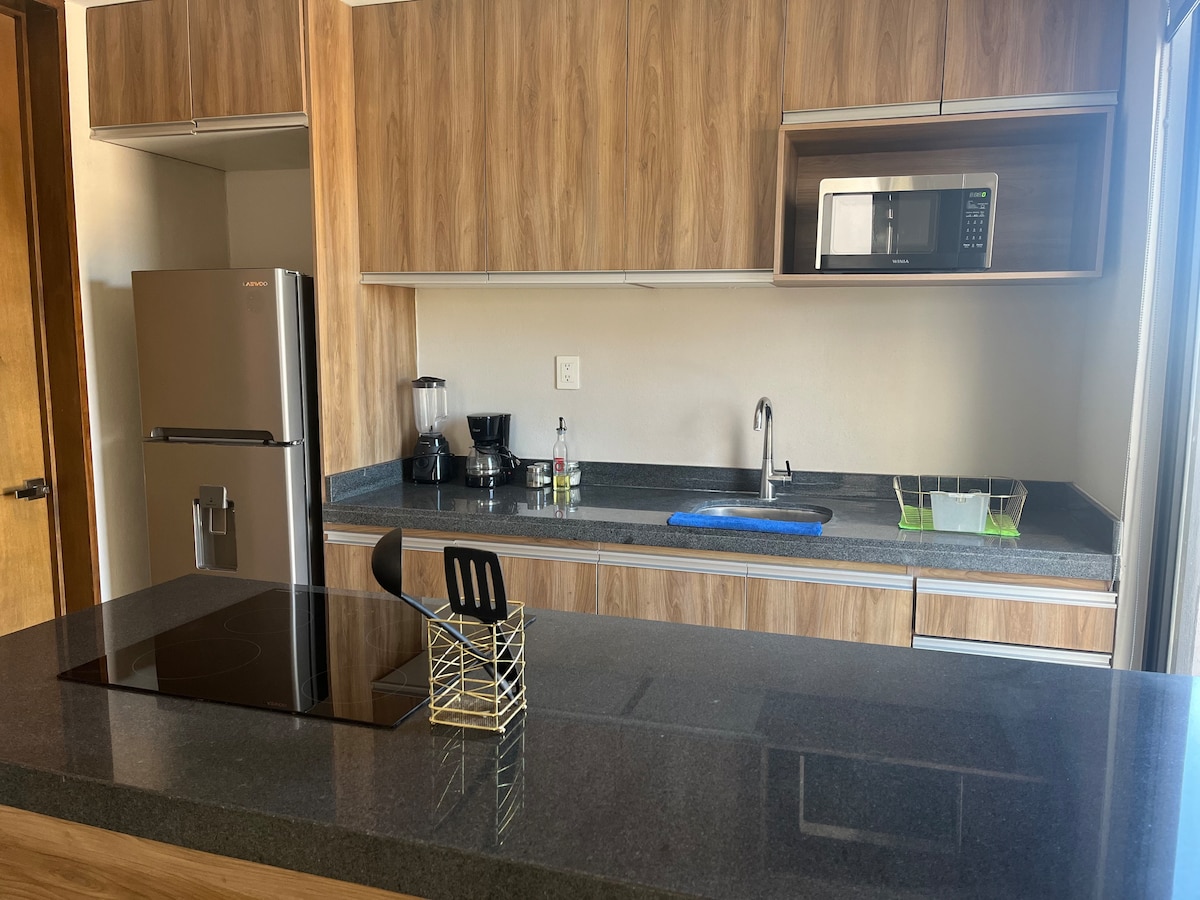 A modern kitchen features wooden cabinetry and a sleek countertop. Stainless steel appliances, including a refrigerator and microwave, are visible. A coffee maker and blender are positioned next to the sink. Natural light filters in through a nearby open window.