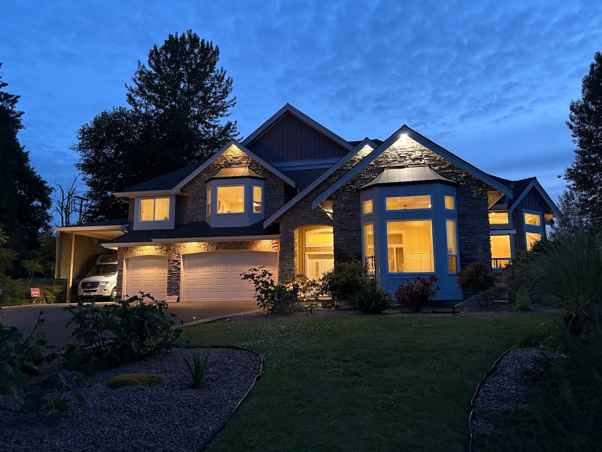 The exterior of a spacious home is shown, illuminated softly at dusk. Large windows reflect warm light, highlighting the stone accents. A well-maintained garden with shrubs and pathways leads to the entrance, while a driveway accommodates vehicle parking. Surrounding trees are visible at the horizon.