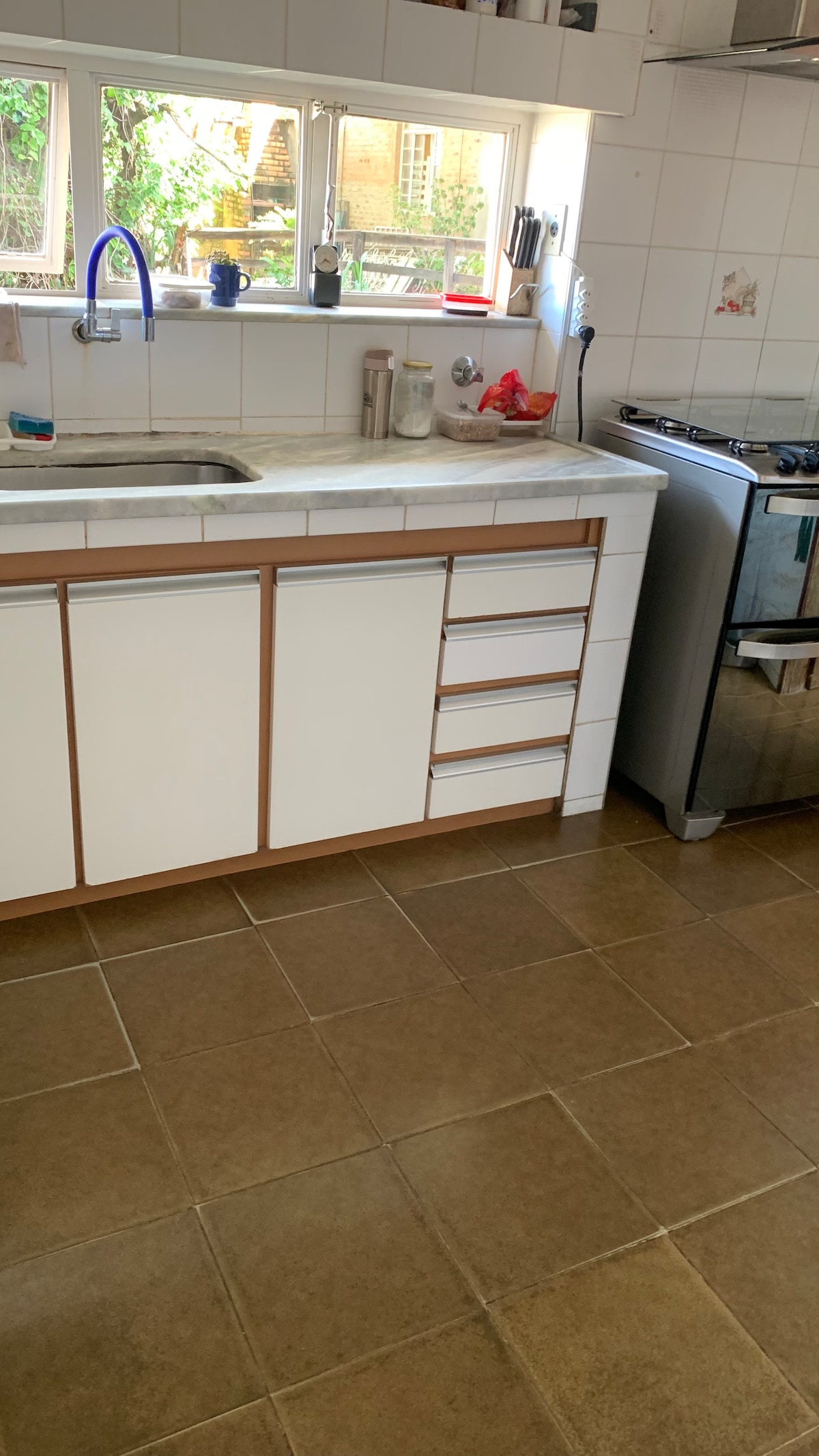 The kitchen area features a white countertop with a sink and several drawers beneath. A stainless steel gas stove is positioned next to the countertop. Large windows provide natural light, and various kitchen items can be seen on the counter and in the windowsill.