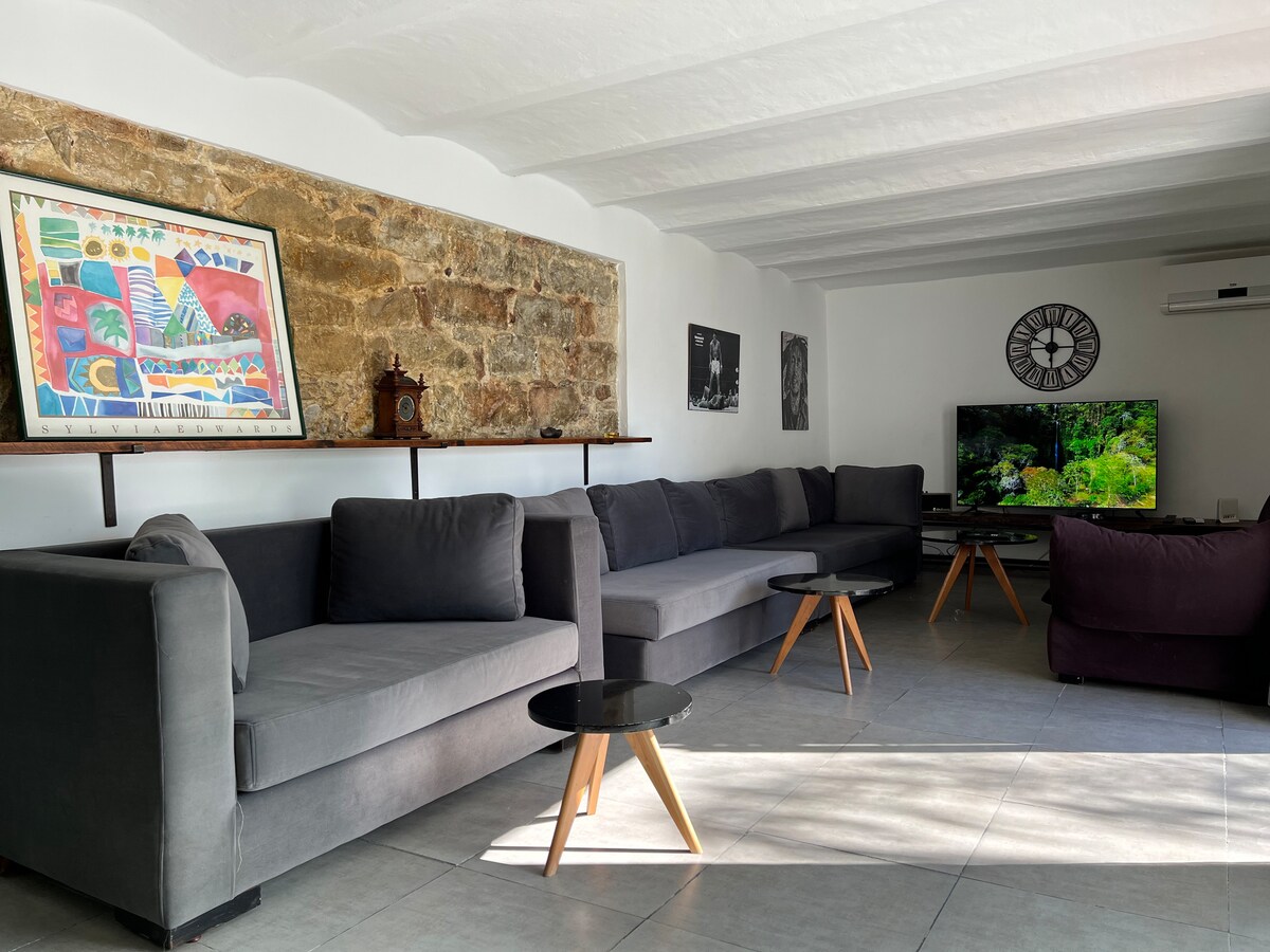 A spacious living area features gray sofas arranged to promote relaxation. A rustic stone wall adds texture, while colorful artwork enhances the ambiance. A low table stands in the center, and a large TV is positioned opposite the seating area, providing entertainment options.