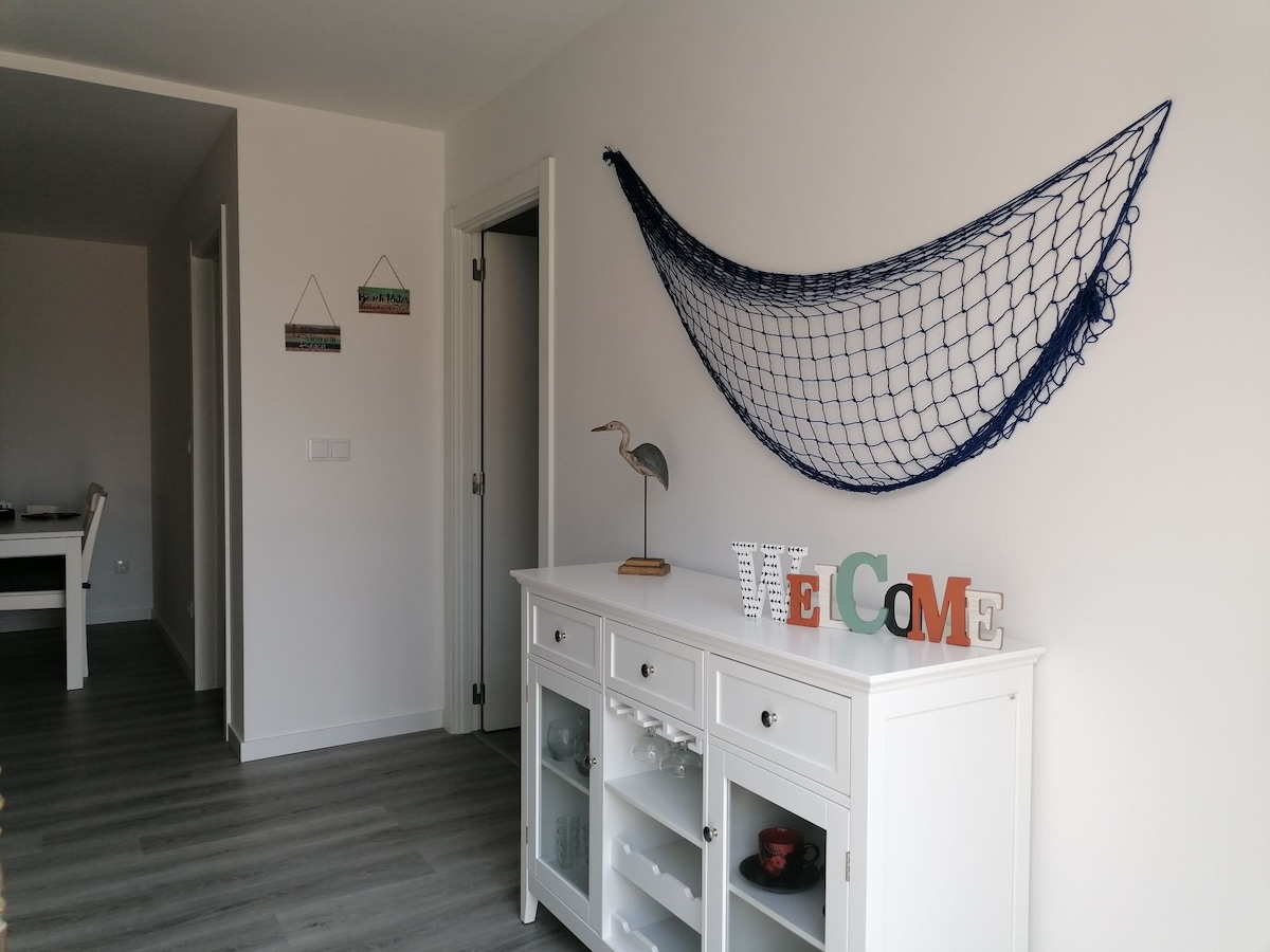 A bright entryway features a white sideboard displaying decorative items, including the word 'WELCOME.' A blue fishing net hangs on the wall above, accompanied by a decorative bird figure. Light-colored walls and flooring create a fresh and open atmosphere.
