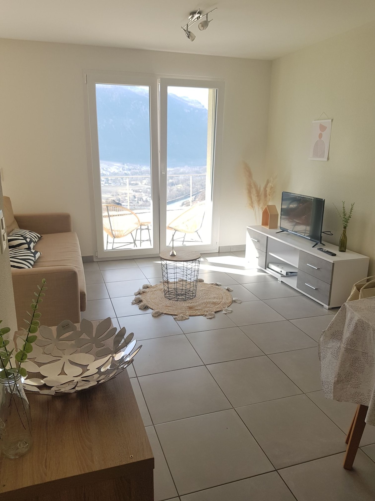 A bright living area features a beige sofa and a flat-screen TV on a light wooden cabinet. Two chairs and a small table are positioned near large glass doors, offering a view of the mountains. Natural light floods the space, highlighted by neutral decor.