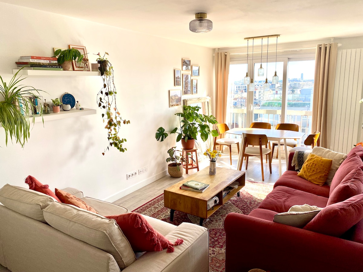 A spacious living area features a mix of cozy seating with a neutral-toned sofa and a vibrant red couch. A wooden coffee table sits in the center, surrounded by greenery, while a dining area with wooden chairs and a table can be seen near large windows revealing a city skyline.