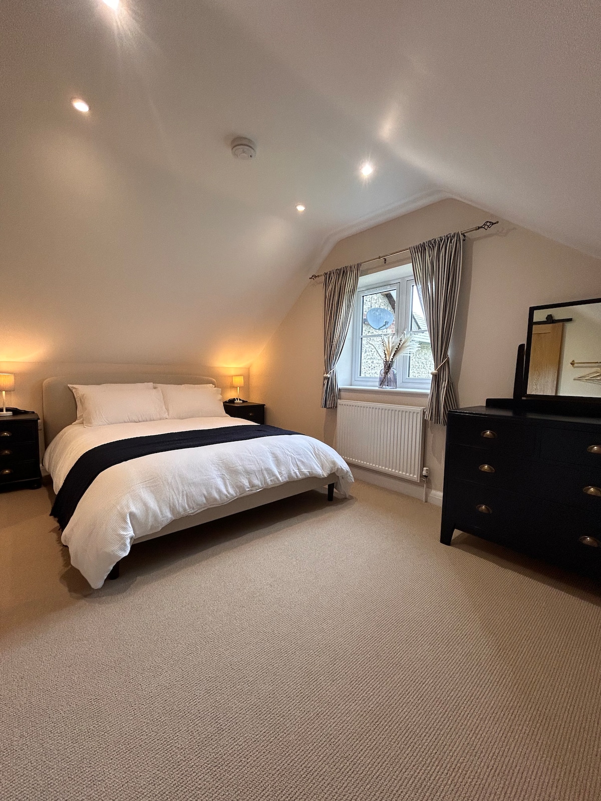 A bedroom features a kingsize bed with a crisp white duvet and a dark blue throw. Soft lighting is provided by two bedside lamps. A window with light curtains allows natural light to enter, and a chest of drawers is positioned near a mirror against the wall.