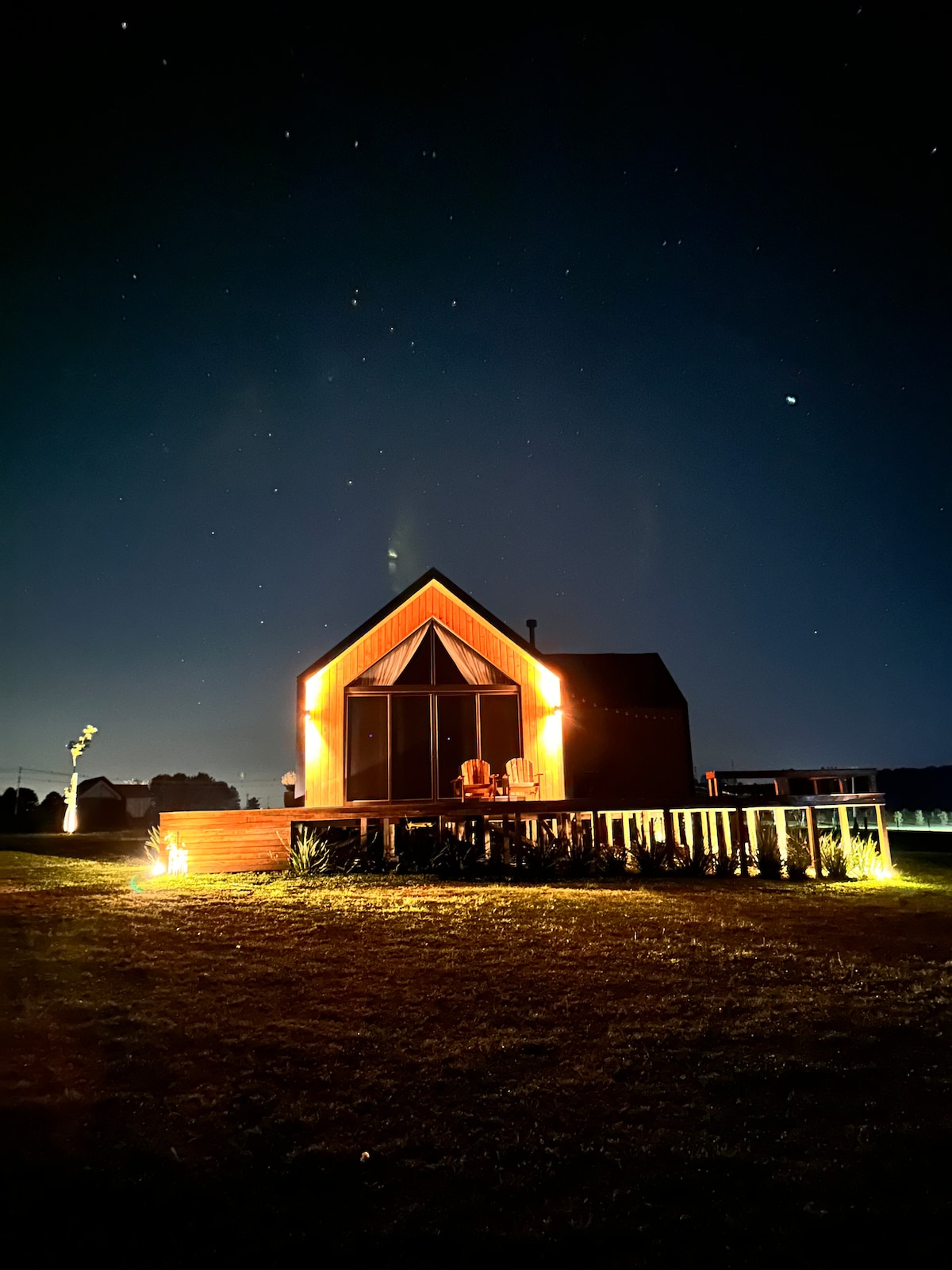 The exterior of a cozy cabin is illuminated against a night sky filled with stars. Warm lights highlight the architectural features, and a porch extends out front, offering seating options. Natural surroundings enhance the tranquil atmosphere of this peaceful retreat.