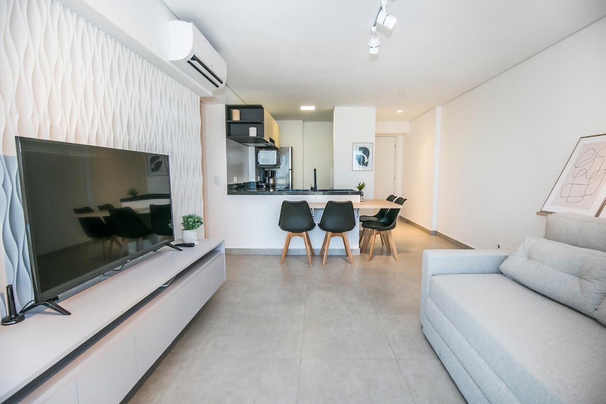 A modern living space is presented, featuring a large flat-screen television and a light-colored sofa. Adjacent is a dining area with a dark wooden table and four chairs. The open kitchen area includes contemporary cabinetry and appliances, enhancing the overall functionality of the room.