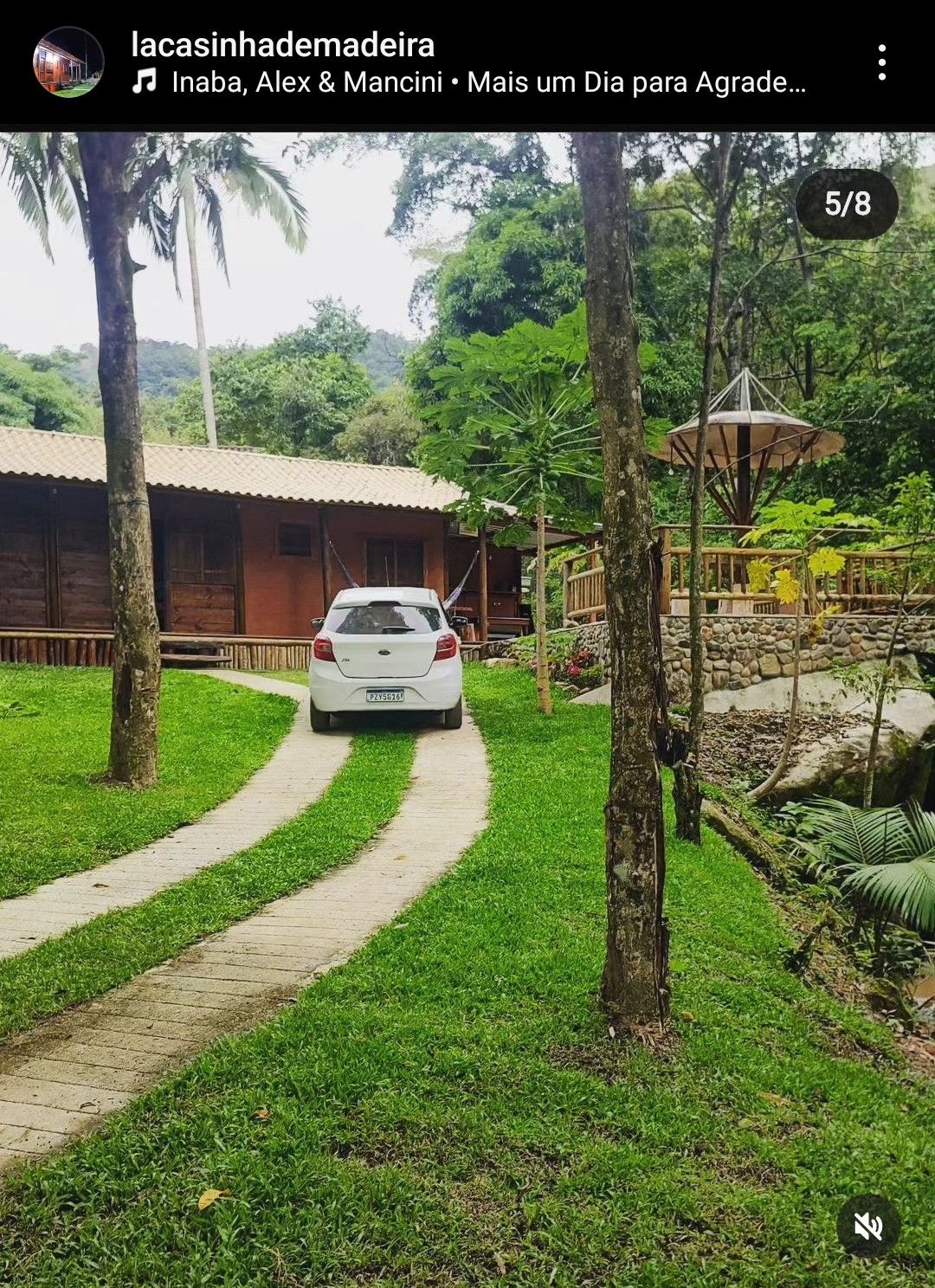 A pathway leads to a charming house nestled among lush greenery. A smooth stone driveway accommodates a parked vehicle. Surrounding trees and plants enhance the natural setting, while nearby, a rustic structure is partially visible, suggesting additional outdoor space.