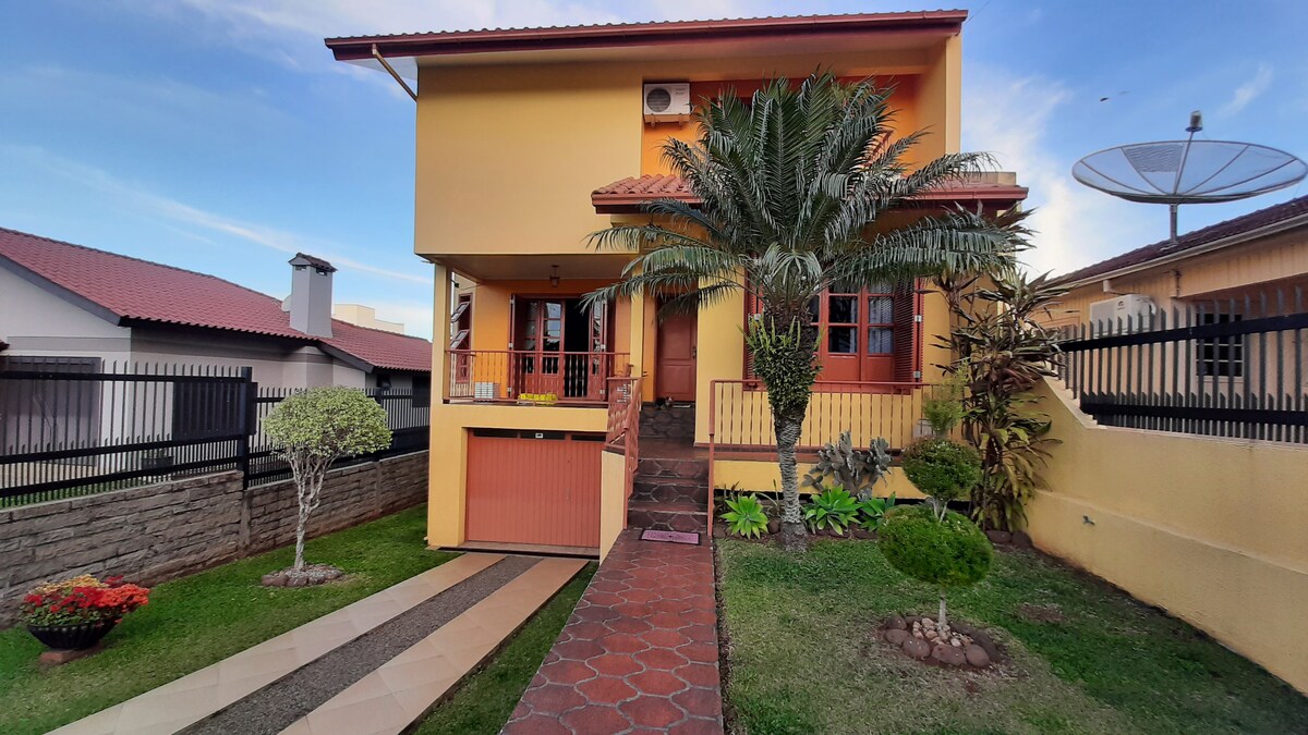 A charming yellow house is surrounded by well-maintained greenery, featuring a pathway leading to the entrance. Palm trees and landscaped shrubs enhance the inviting front yard. A satellite dish is visible on the roof, indicating modern amenities, while the fenced property offers privacy.