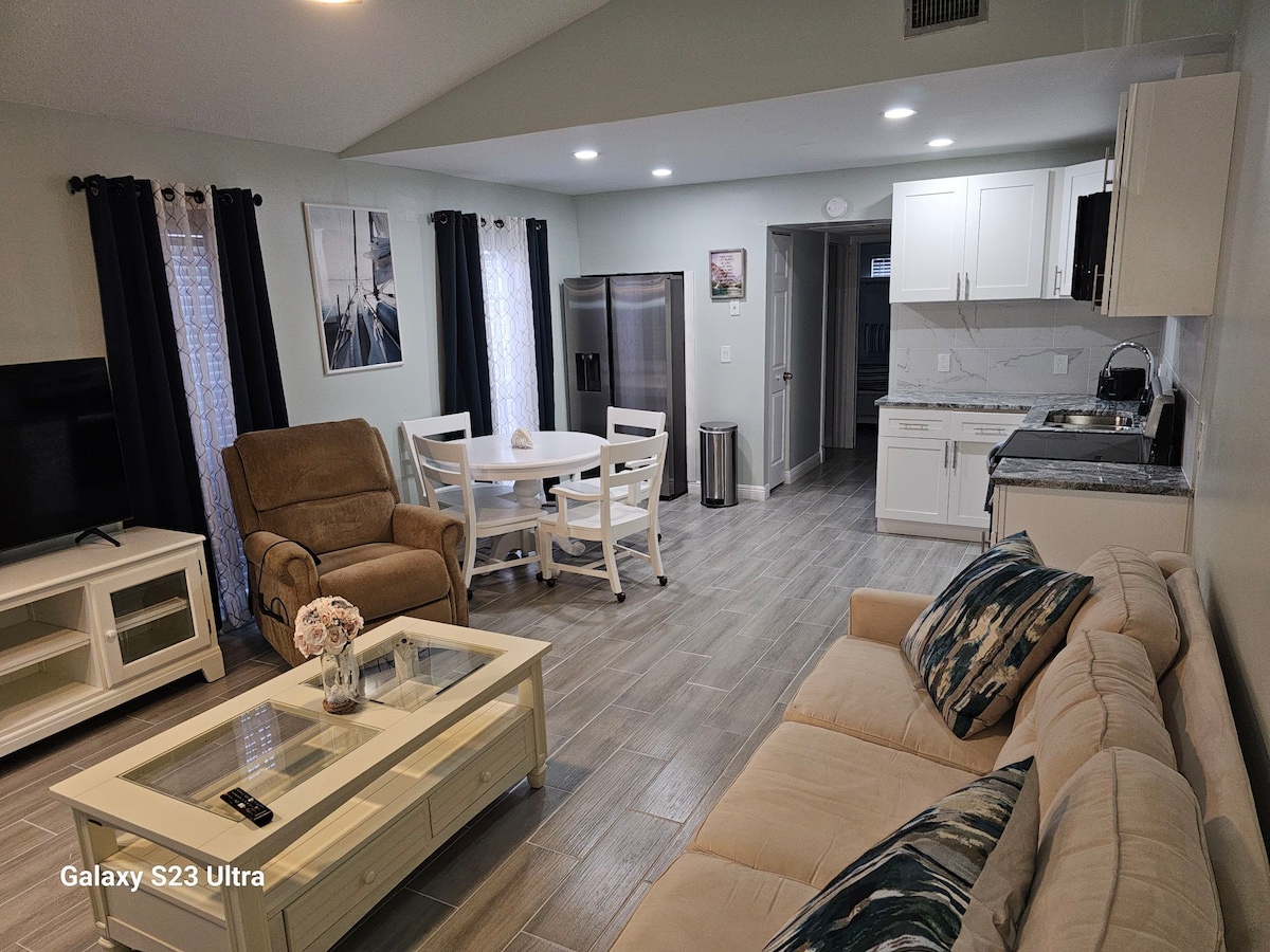 An open living space includes a comfortable light-colored sofa adorned with decorative pillows. A dining table with four chairs is positioned nearby, and a kitchenette with modern appliances is visible in the background. Large windows allow natural light to fill the room.