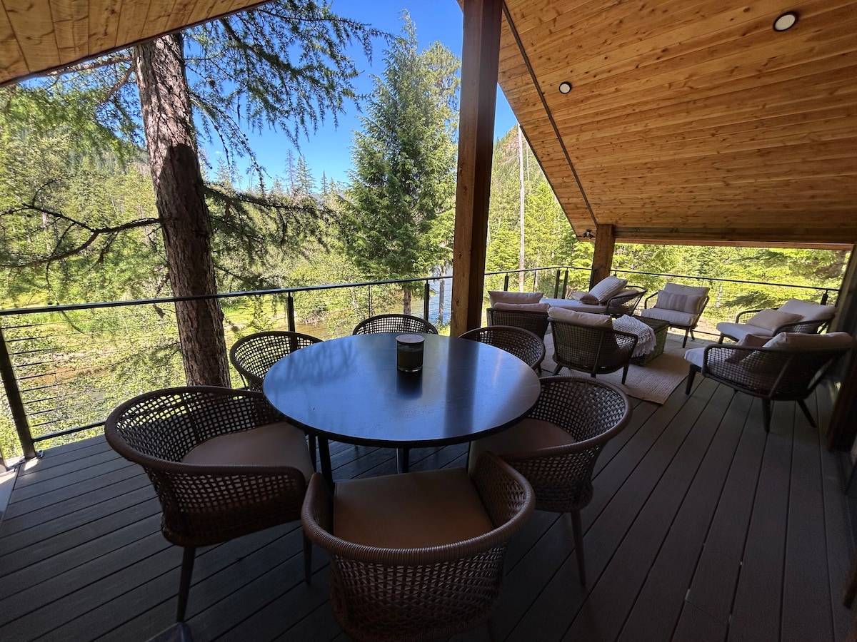 A covered deck area is presented with a round dining table surrounded by woven chairs. Sunlight filters through the trees, enhancing the natural scenery. A lounge area with plush seating is visible in the background, inviting relaxation amid the tranquil forest surroundings.