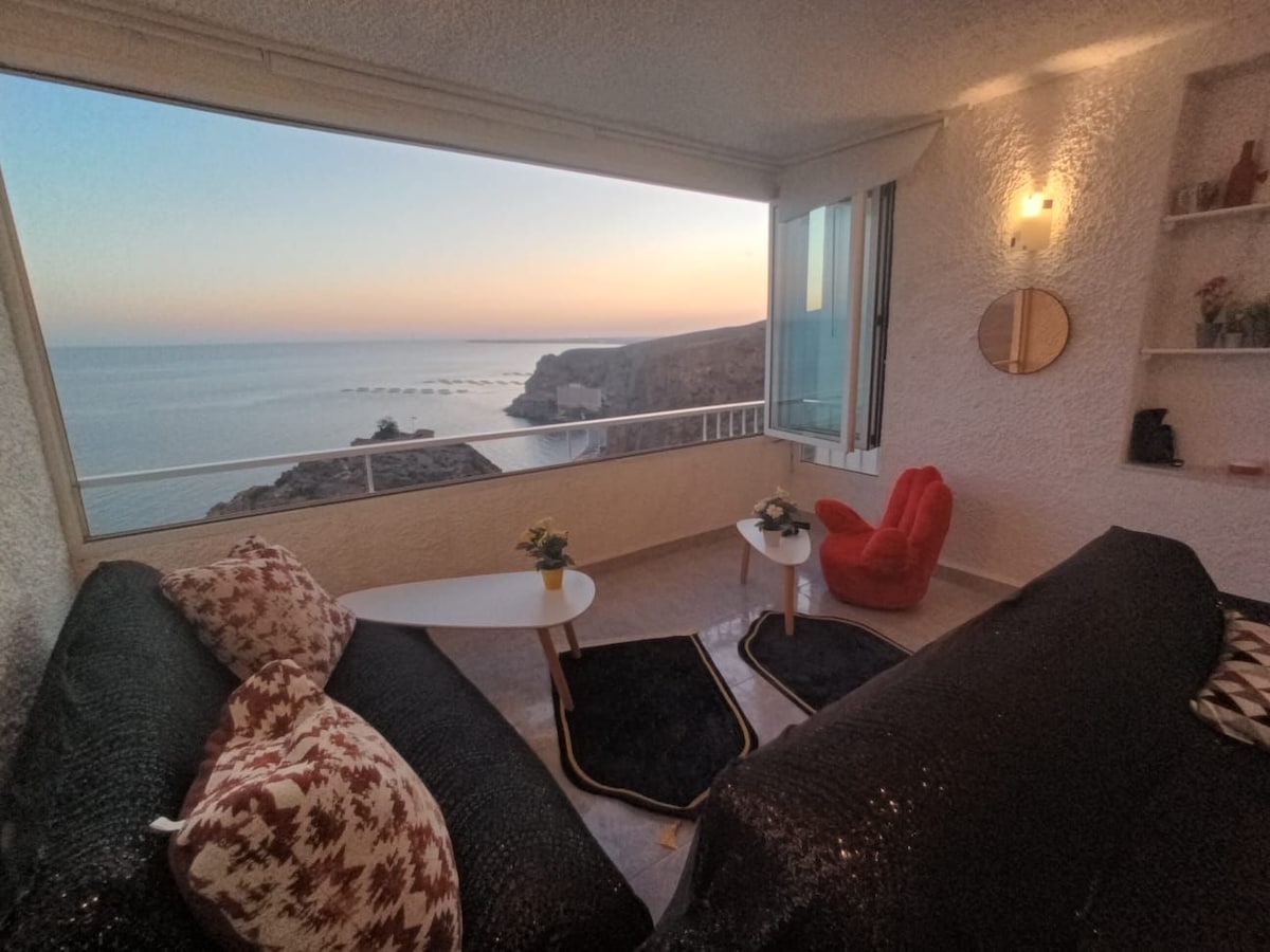 A cozy seating area is highlighted by large windows offering expansive ocean views at sunset. Two black sofas, accentuated with decorative pillows, face a small table. A vibrant red chair adds a pop of color to the space, creating an inviting atmosphere for relaxation.